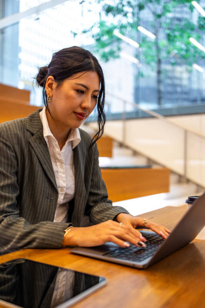istock Young professional Asian woman working on a tablet and laptop in a modern office setting, showcasing business technology and productivity. The image captures the professional work environment and focus on multitasking in an urban setting. 2158070359