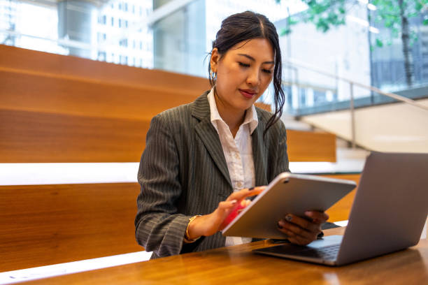 Young professional Asian woman working on a tablet and laptop in a modern office setting, showcasing business technology and productivity. The image captures the professional work environment and focus on multitasking in an urban setting.