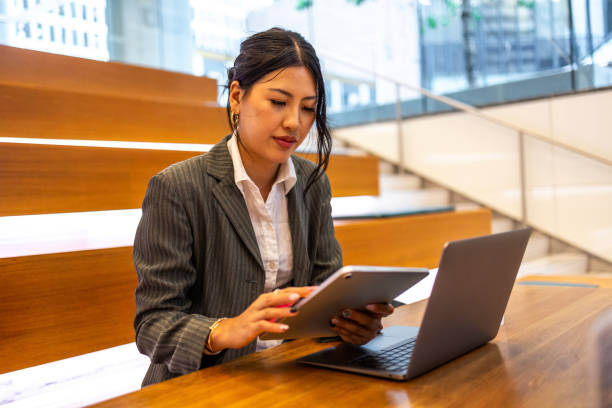 istock Young professional Asian woman working on a tablet and laptop in a modern office setting, showcasing business technology and productivity. The image captures the professional work environment and focus on multitasking in an urban setting. 2158070361