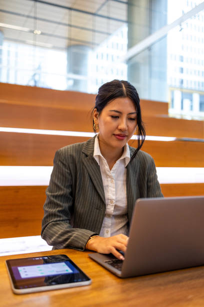 istock Young professional Asian woman working on a tablet and laptop in a modern office setting, showcasing business technology and productivity. The image captures the professional work environment and focus on multitasking in an urban setting. 2158070365