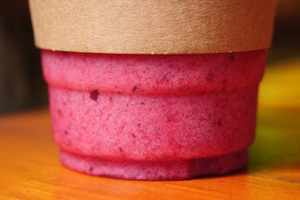 A close-up shot of a red berry smoothie in a plastic cup on a wooden surface. The smoothie is a vibrant pink red color and has a smooth, creamy texture.