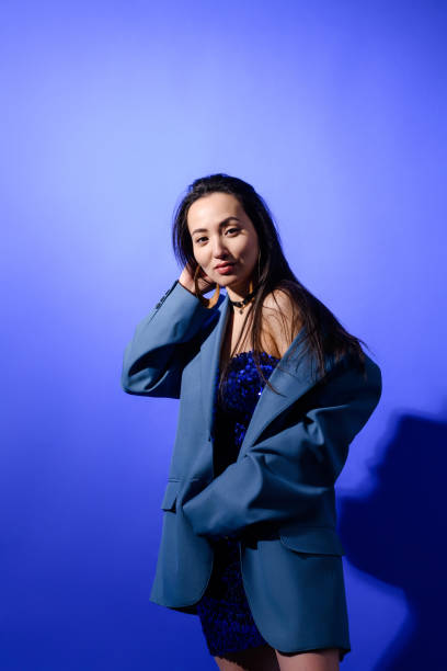 istock Trendy asian model in jacket and dress looking at camera on blue background 2158094625