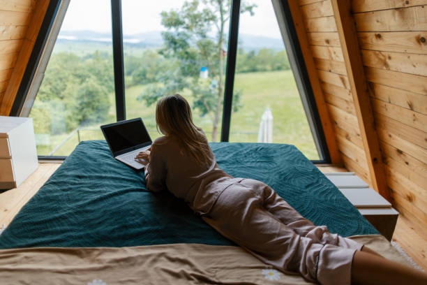 Beautiful mid adult woman working on her laptop at home in mountain house. She is in her pajamas. lying on the bed.