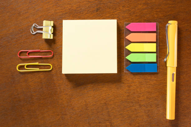 A yellow pen sits on a table with a stack of colorful sticky notes and a pair of red and yellow paper clips.