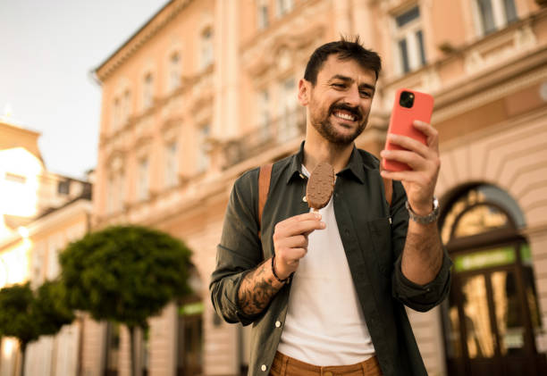 istock Happy young man using phone and eating Ice Cream while exploring the city. 2158125359