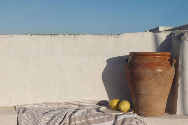 Vintage olive clay storage pot, vase with fresh lemon fruit and textile throw, blanket. White old textured white wall mockup in sunlight. Blue sky background, summer still life, breakfast composition.
