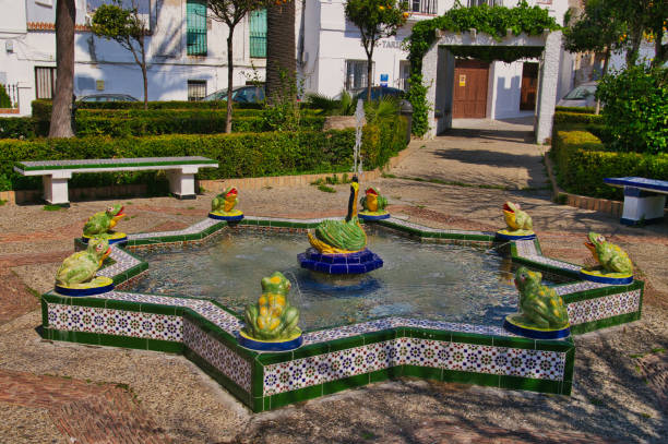 Star-shaped fountain with ceramic green frogs and a yellow and green swan in the middle. Background of green hedges and white houses.