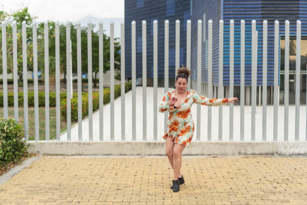 Woman dancing outdoors in a floral dress near a metal fence and modern building.