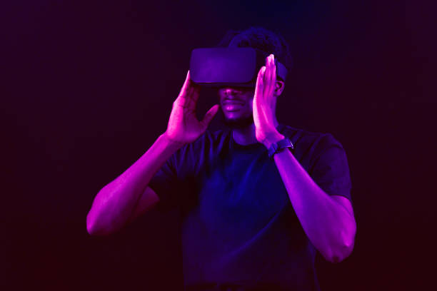 istock Young african metaverse user wearing virtual reality headset, playing new VR game online, touching something invisible in neon light. Metaverse technology concept. African man with VR virtual reality goggles. Futuristic lifestyle. 2158154297