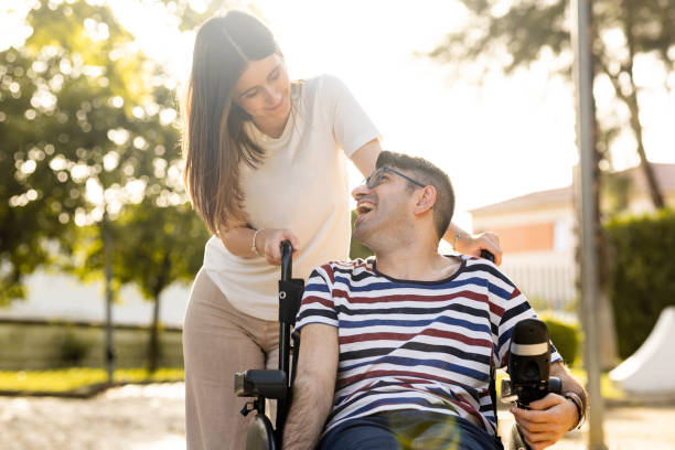 un hombre adulto con una discapacidad se sienta en una silla de ruedas automática con una mujer joven en un parque al atardecer. la mujer y el hombre se miran felices. concepto de apoyo a las personas con discapacidad. - adulto fotografías e imágenes de stock