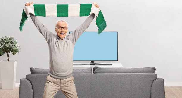 istock Elderly man cheering with a scarf in front of a sofa 2158182218