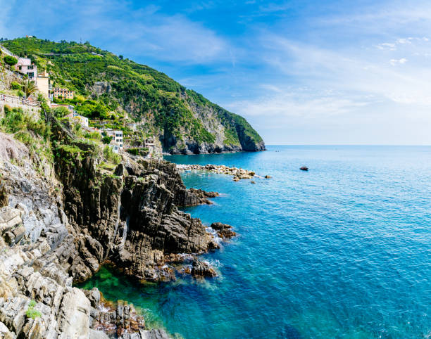 Coastal cliffs descend into crystal clear water with rocky outcrops visible. The rocky cliffs are covered in green vegetation, adding natural beauty to the rugged landscape. The clear water showcases underwater rocks and the diverse marine environment along the coast.