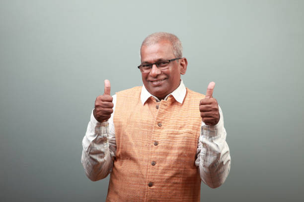 A South Indian old man with a bold expression says super in front of the camera. His finger is out of focus in the foreground, creating a sense of depth. The background is a plain white wall.