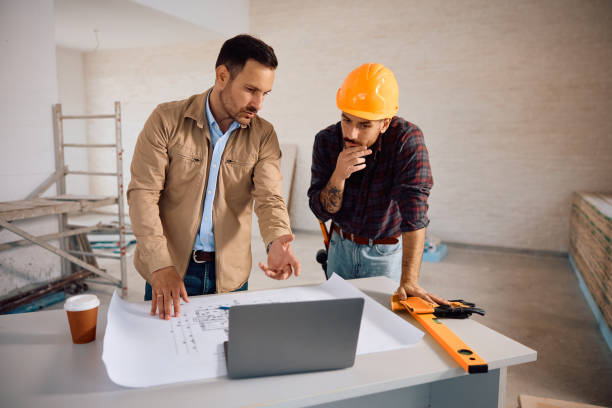istock Mid adult man and construction worker using laptop while analyzing blueprints at renovating home. 2158230752