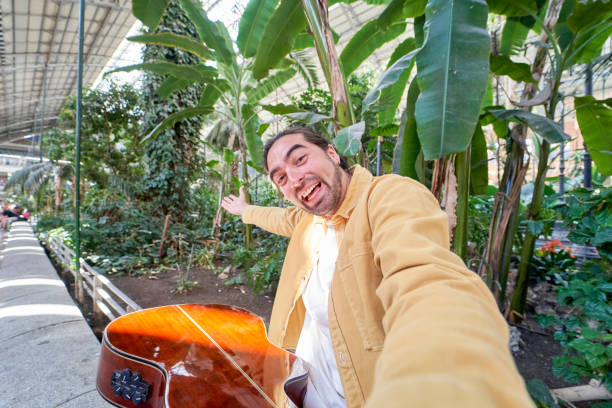 man sitting on a bench with large green plants in the background taking a selfie with his smartphone