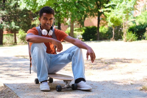 A young student man is sitting on a skateboard in a park. He is wearing headphones and a backpack