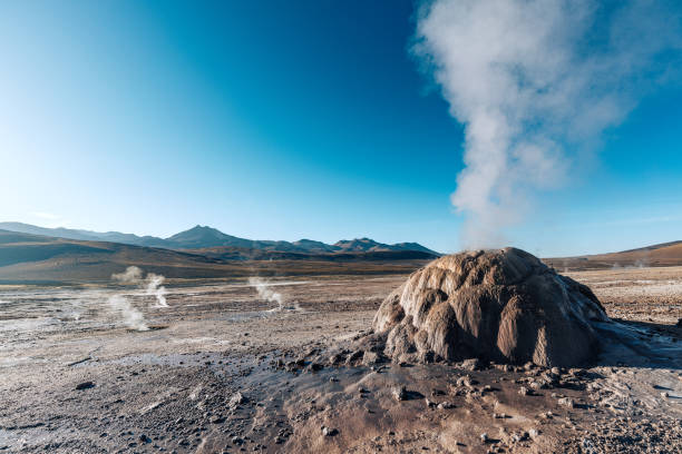 Steam rises from a geothermal formation in the Atacama Desert, Chile, under a clear blue sky with mountains in the background.