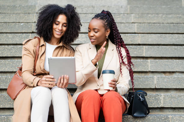 istock Two Black Businesswomen Discussing Business Ideas on Tablet While Sitting on Steps 2158245135