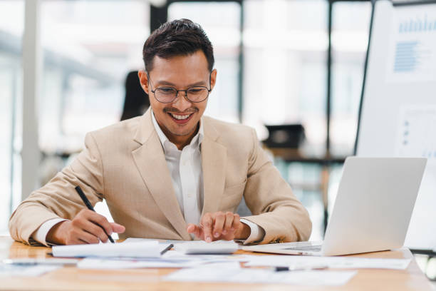 istock A handsome, smiling businessman wearing glasses and a suit, sitting at a desk while working on a laptop. 2158254636
