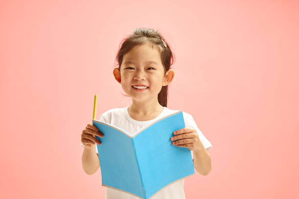istock Joyful First Grader Girl Reader with Exercise Book Against Pink Isolated 2158261841
