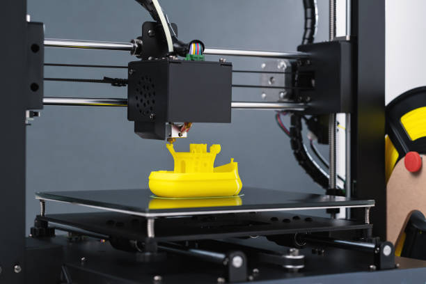 istock Printing on 3D printer figurines toys ship from yellow plastic close-up. Clear movements of the print head and printer working platform. Potential of modern technology 3D printing. Plastic reel 2158263274