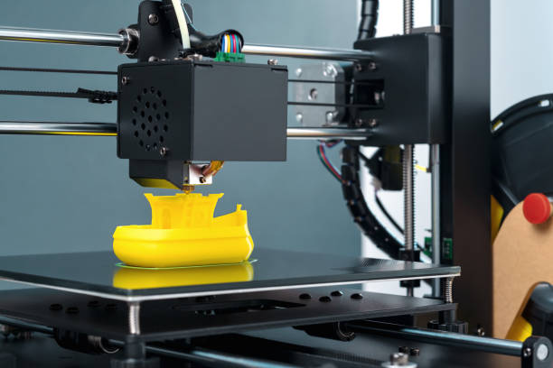 istock Printing on 3D printer figurines toys ship from yellow plastic close-up. Clear movements of the print head and printer working platform. Potential of modern technology 3D printing. Plastic reel 2158263578