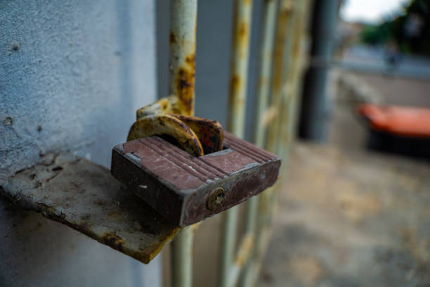 A close-up photo of a rusty padlock securing a metal gate. The padlock is brown with a visible keyhole and is attached to a metal bar that is also rusty. The gate is made of metal bars and is painted a light gray color. The background is blurry and shows a sidewalk and a green plant.