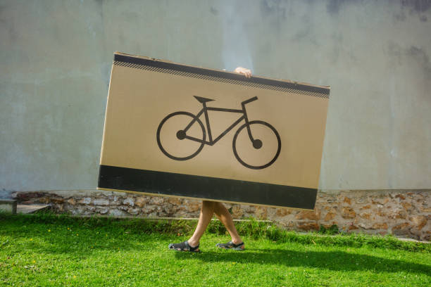 Person hide and carrying an enormous box marked with a bike logo Person hidden behind a sizable box featuring a bike illustration, walking on grass with just their lower limbs visible legs against the wall stock pictures, royalty-free photos & images