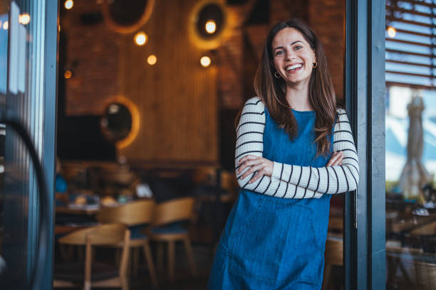 Smiling Female Barista in Coffee Shop A joyful woman in a striped shirt and denim apron stands with arms crossed at a welcoming coffee house, embodying professional hospitality. small business stock pictures, royalty-free photos & images