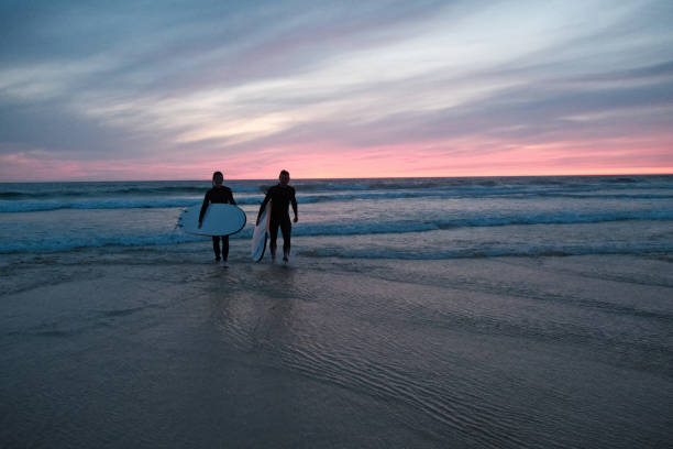 Couple with stand up paddle boards walk up beach at sunset