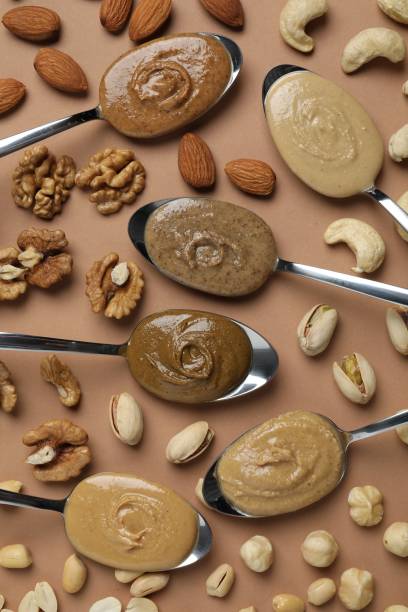 Tasty nut butters in spoons and raw nuts on light brown table, flat lay