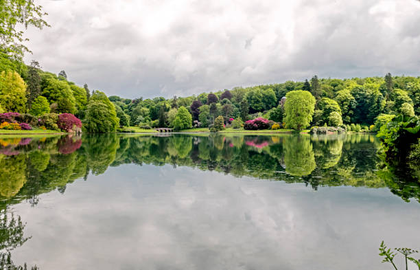 Crisp mirror reflection in a still lake, of a classic old stone English Bridge. The bridge is in the distance, connecting the beautiful horizon of trees, green foliage and pink rhododendron flowers from the English countryside where visitors take pleasant lakeside walks. A sunny day with fluffy clouds in the sky and reflected in the lake gives room for copy space.