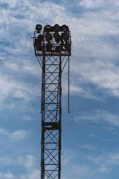 istock A tall industrial lattice tower against a clear blue sky with clouds. Several large round lights, similar to those used to illuminate stadiums or sports grounds, are mounted on the tower. 2158334943