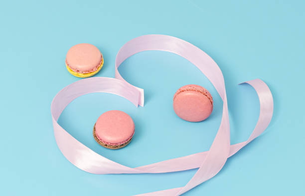 Three colorful macaroons and a twisted heart-shaped pink ribbon on a pale blue background, close-up side view.Sweet dessert concept.