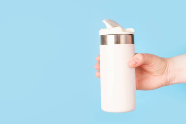 istock Female hand holding white thermos mug or thermos on blue background side view. 2158344539