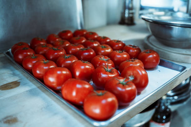 A sheet of ripe, red tomatoes sit on the counter of an industrial kitchen, waiting to be used to cook with.