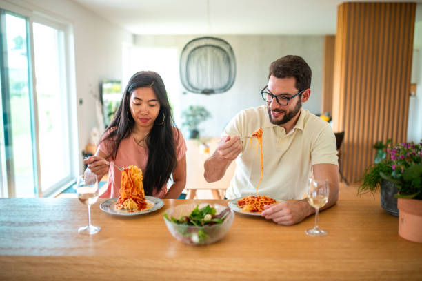 An Asian woman and a Hispanic man enjoy a savory pasta dish, sharing a meal together at a well-appointed domestic dining table.