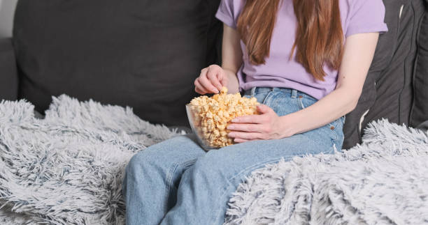 istock Lonely, unrecognizable woman, sitting on couch eating popcorn from glass plate while watching TV 2158379103