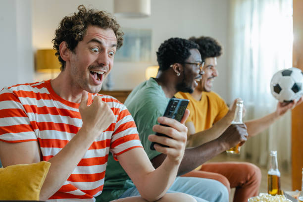 istock Cheering and winning. A young man is holding a mobile phone while watching sports with his friends at home 2158383304