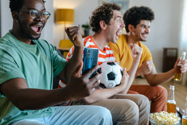 istock Cheering and winning. African American young man is holding a mobile phone while watching sports with his friends at home 2158383320
