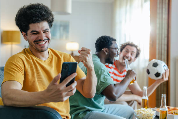 istock Cheering and winning. A young man is holding a mobile phone while watching sports with his friends at home 2158383594