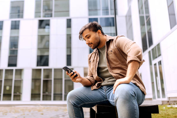 istock Casually clothed man using smart phone while sitting on a bench in front of modern building 2158384105
