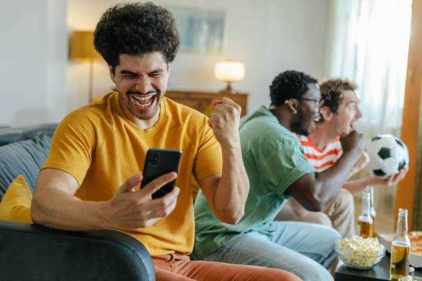 istock Cheering and winning. A young man is holding a mobile phone while watching sports with his friends at home 2158384160