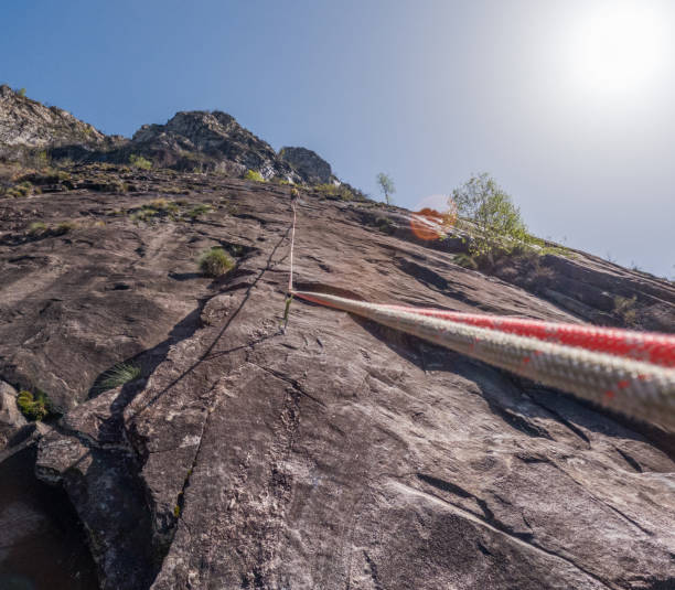 View from below of a climber ascending a steep rock face using a climbing rope, highlighting the thrill and challenge of rock climbing under the bright sun