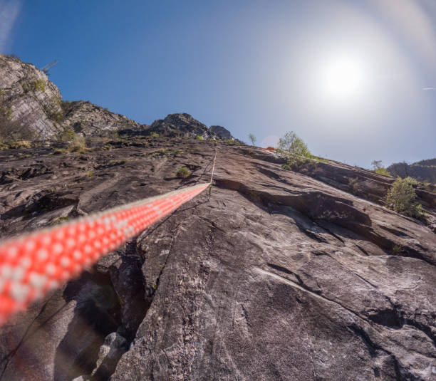 View from below of a climber ascending a steep rock face using a climbing rope, highlighting the thrill and challenge of rock climbing under the bright sun