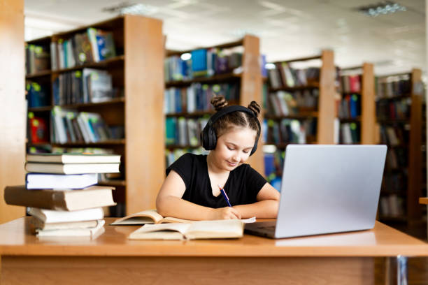istock School library. Schoolgirl studying online using laptop, taking notes on paper. Teenage schoolgirl wearing headphones undergoes online learning. Distance learning concept. 2158400598
