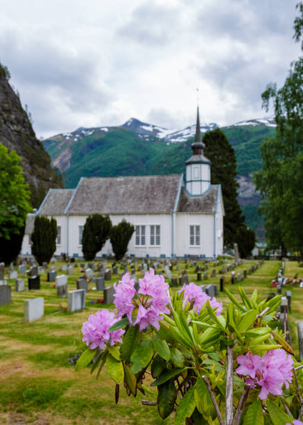 A white church stands against a backdrop of snow-capped mountains in Norway, surrounded by a graveyard filled with colorful flowers. Valldal Norway