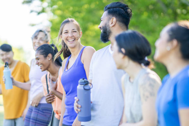 A large group of mature adults are seen standing together outside after a fitness class.  They are each dressed comfortably in athletic wear and are hydrating as they talk amongst themselves.