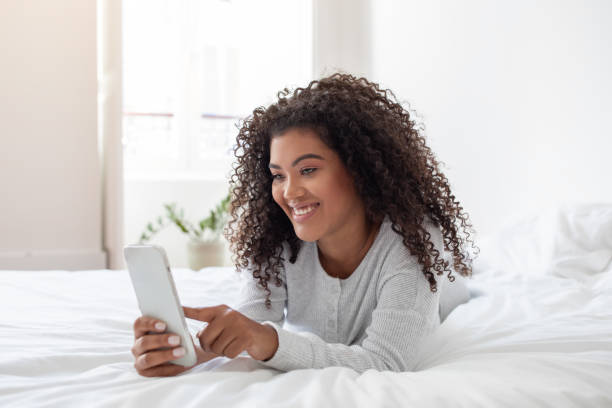 istock Young Woman Smiling While Using Phone in Bedroom During Morning 2158420581