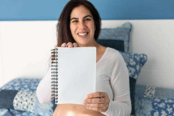 Delighted pregnant female sitting on soft bed at home and showing blank notepad at camera